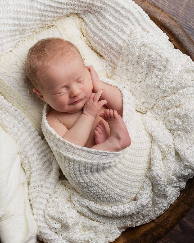 Newborn baby girl smiles softly while bundled up in white fabric for her newborn session in a Davison studio.