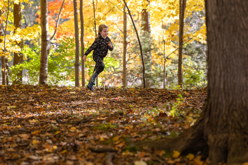 A young boy is running through the woods over crunchy leaves during his fall family photo session at Flushing County Park in Genesee County.